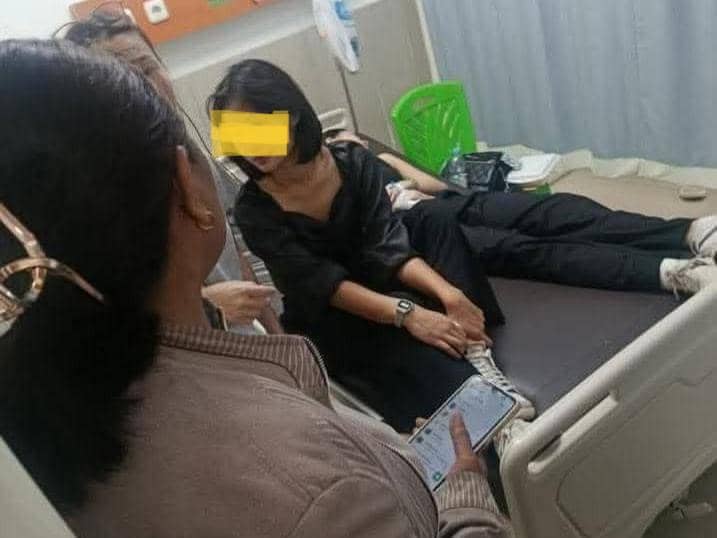 Seorang perempuan duduk di tepi ranjang rumah sakit bersama temannya yang terbaring, didampingi dua orang lainnya di ruang perawatan.