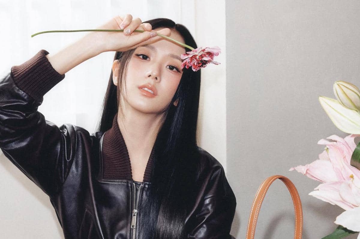 Seorang wanita berambut panjang mengenakan jaket kulit hitam memegang bunga merah muda di depan wajahnya di dalam ruangan.