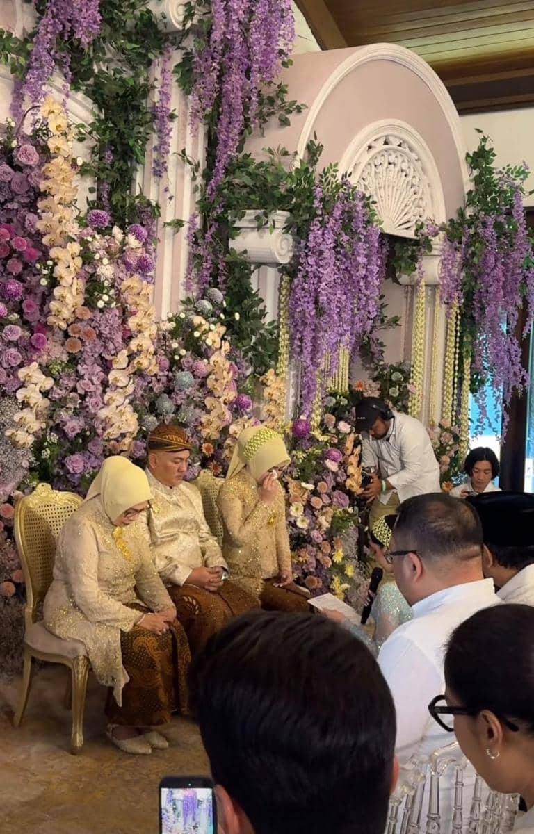 Suasana pengajian dan siraman dengan keluarga berpakaian adat warna emas duduk di pelaminan berhias bunga ungu dan krem.