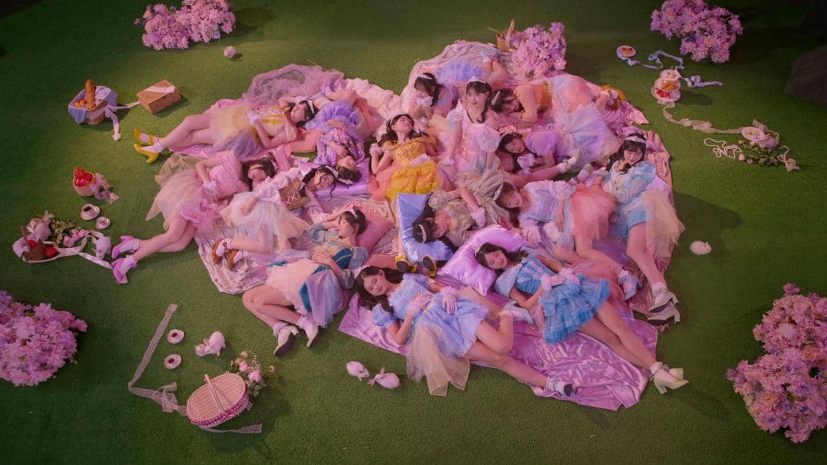 Belasan anggota grup idola mengenakan gaun warna pastel berpose berbaring di atas kain ungu berbentuk hati dengan latar rumput hijau dan bunga sakura.