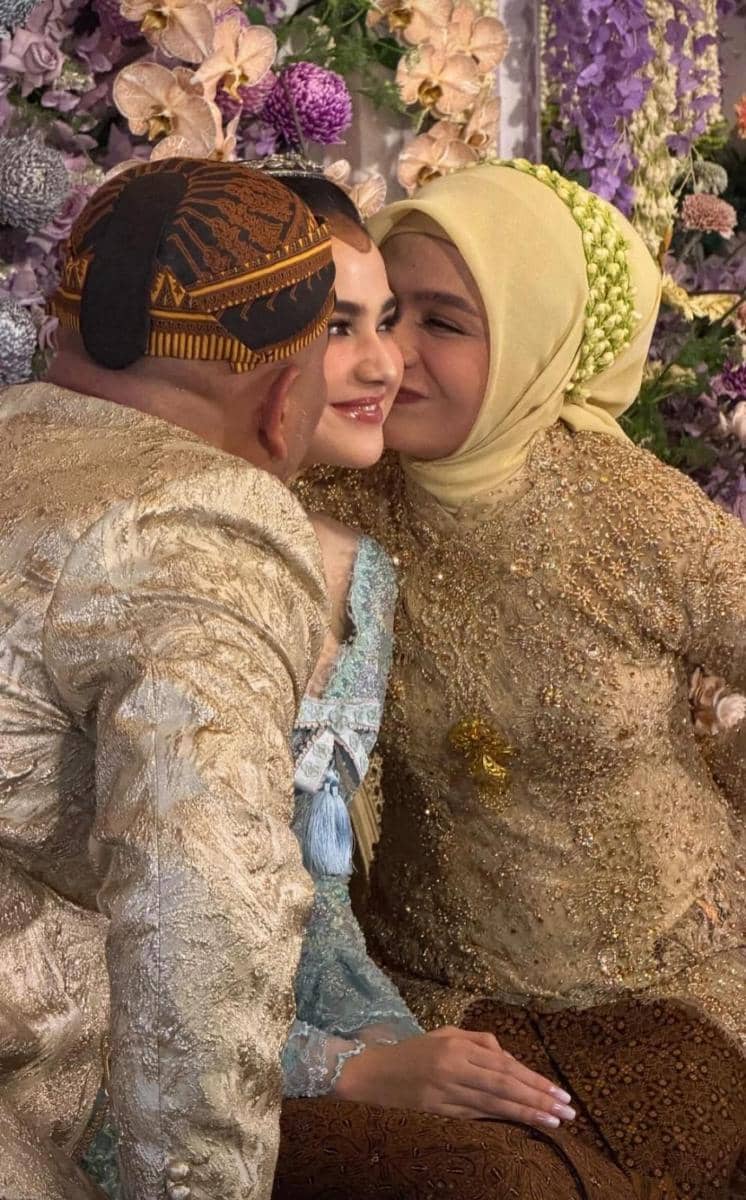 Seorang perempuan berbusana kebaya biru duduk diapit dua orang tua berpakaian adat Jawa berwarna keemasan dalam suasana pengajian dan siraman.