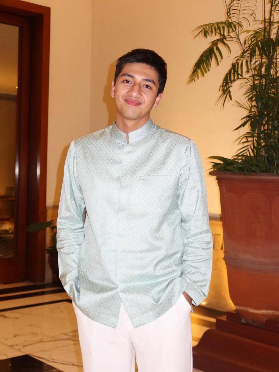 Harris Vriza mengenakan baju koko berwarna hijau muda dan celana putih berdiri di dalam ruangan dengan pot tanaman besar di belakangnya.