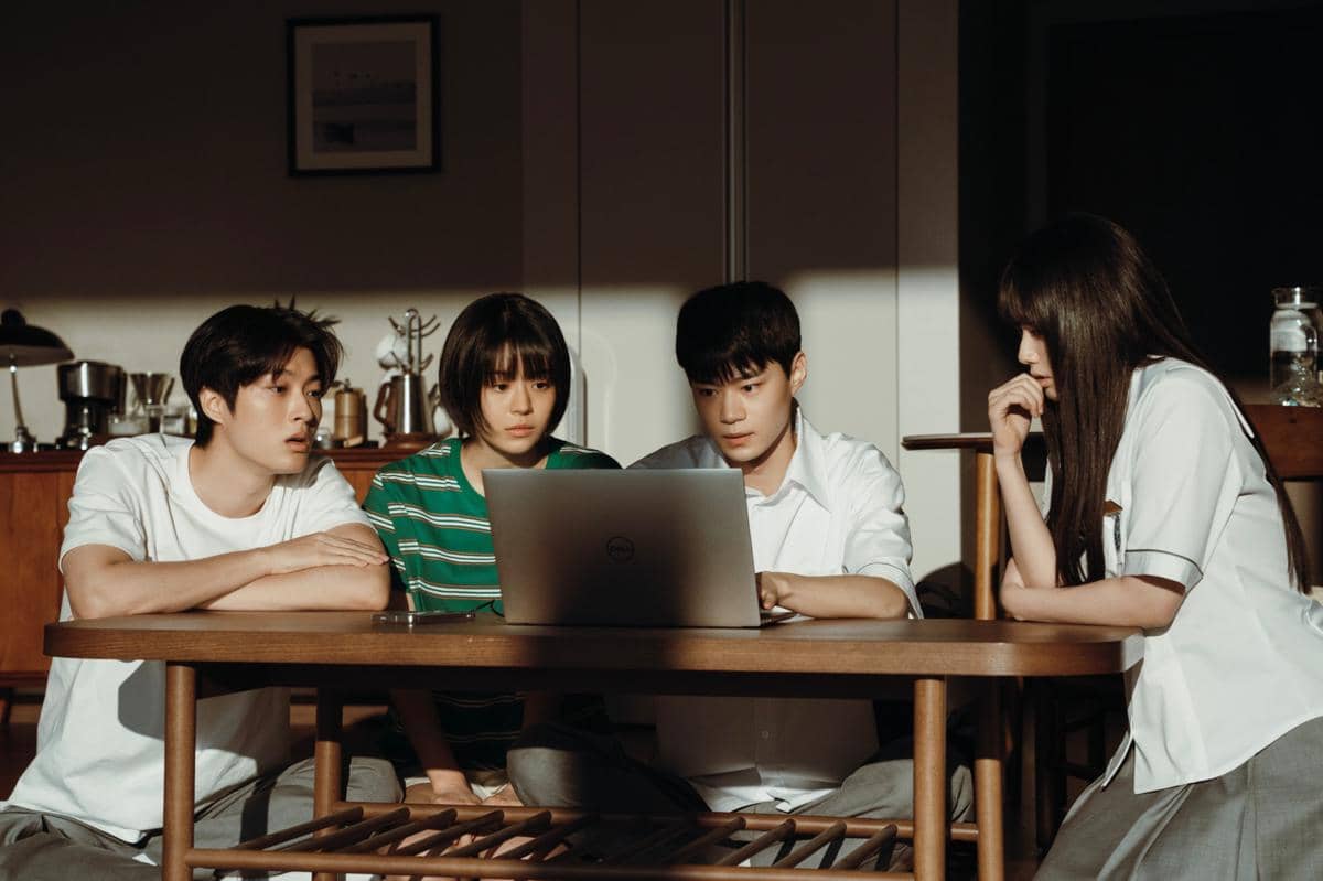 Empat remaja duduk di sekitar meja kayu sambil menatap layar laptop dengan ekspresi serius dalam adegan drama Korea If Wishes Could Kill.