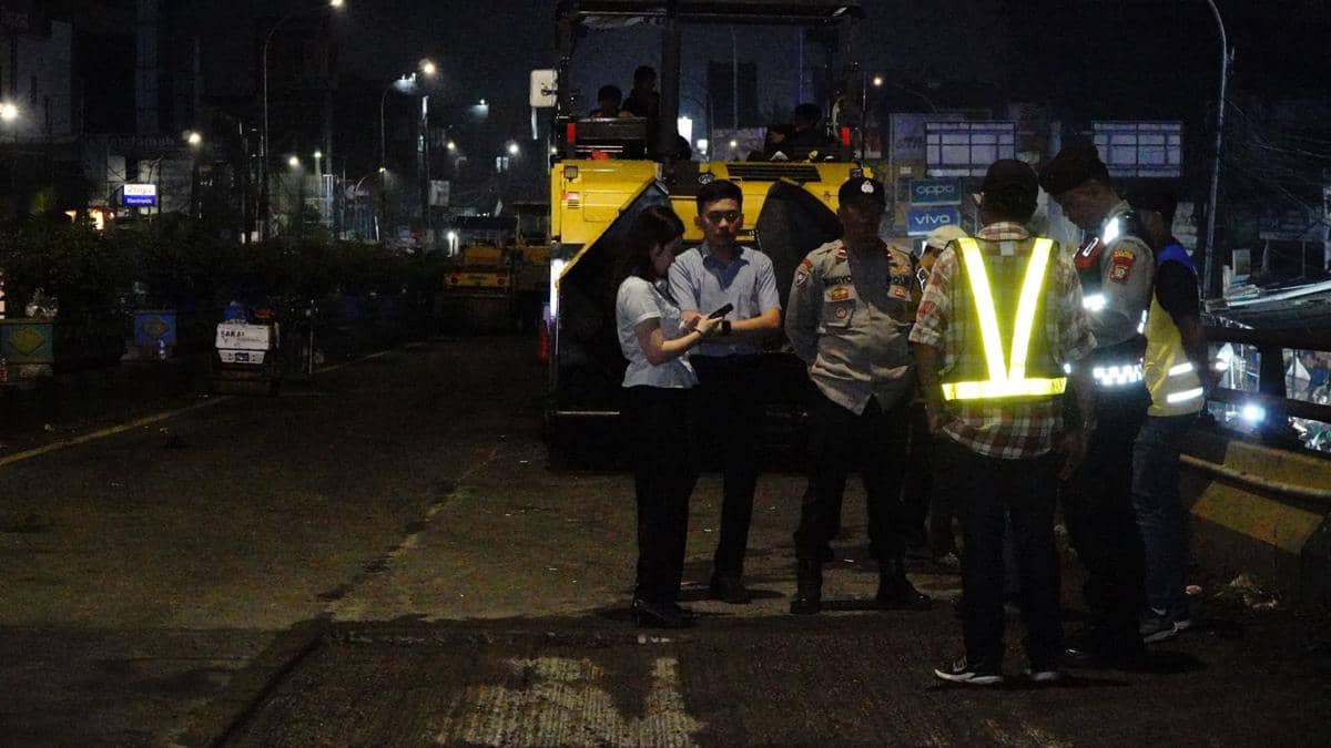 Beberapa petugas dan pekerja berdiskusi di lokasi proyek perbaikan Flyover Ciputat pada malam hari dengan alat berat di belakang mereka.