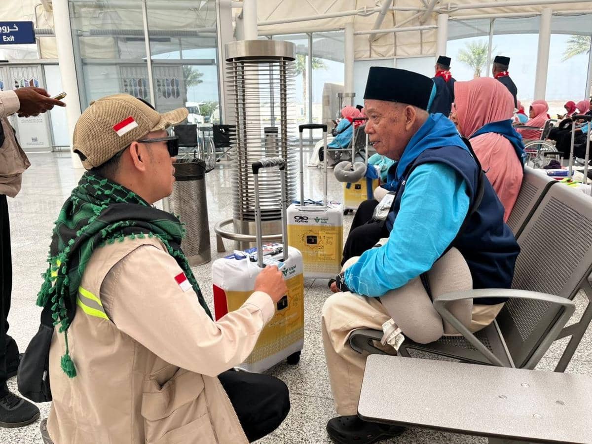 Petugas Haji berbicara dengan jamaah lanjut usia di ruang tunggu bandara, memberikan perhatian dan bantuan sebelum keberangkatan.