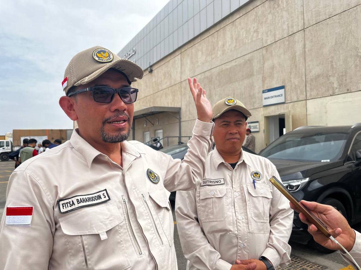 Dua petugas berpakaian seragam krem dengan topi dan kacamata berdiri di area parkir dekat gedung bandara, salah satunya berbicara.