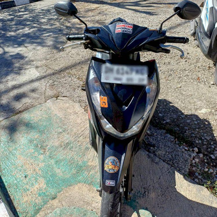 Sebuah sepeda motor hitam dengan stiker di bagian depan terparkir di area beraspal di Kota Kupang pada siang hari.