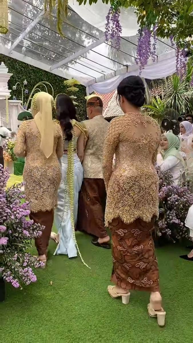 Beberapa orang mengenakan kebaya dan kain batik berjalan di taman berhias bunga ungu saat acara pengajian dan siraman.