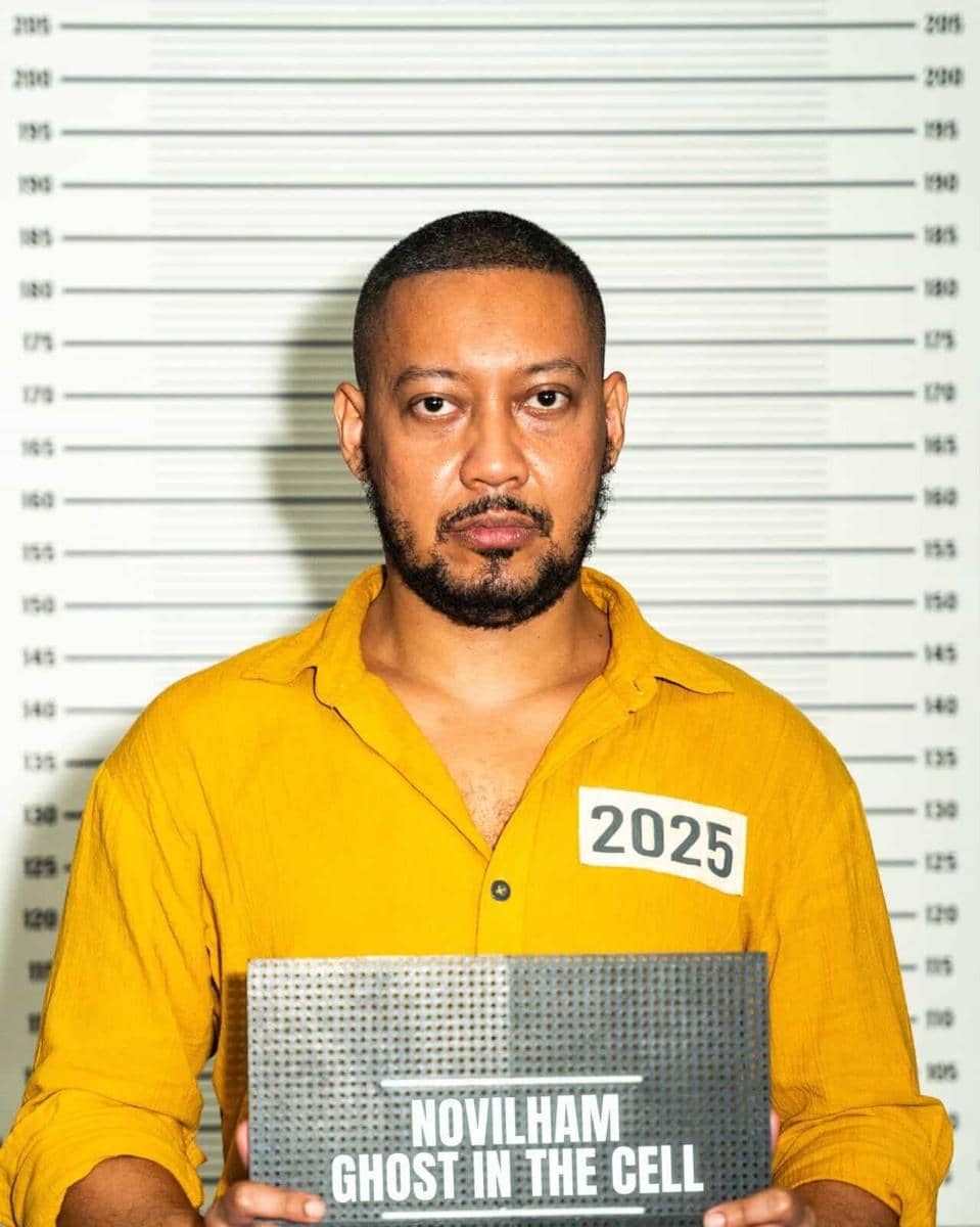 Seorang pria mengenakan kemeja kuning dengan label angka 2025 memegang papan bertuliskan 'Novilham Ghost in the Cell' di depan latar tinggi badan.