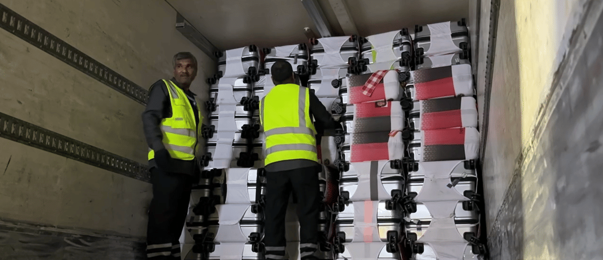 Dua porter bandara mengenakan rompi keselamatan kuning sedang menyusun koper di dalam truk pengangkut bagasi.