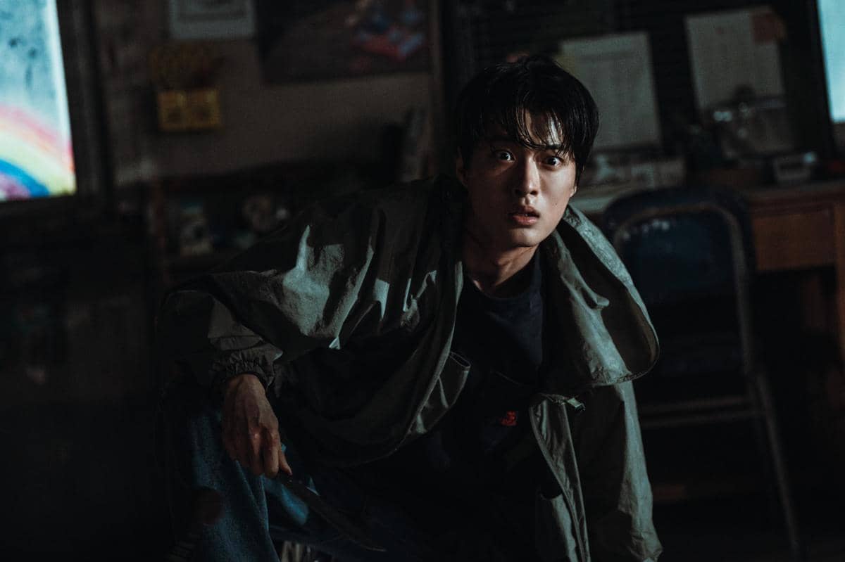 Seorang pria dengan ekspresi tegang berjongkok di ruangan gelap sambil memegang benda tajam dalam adegan drama Korea If Wishes Could Kill.