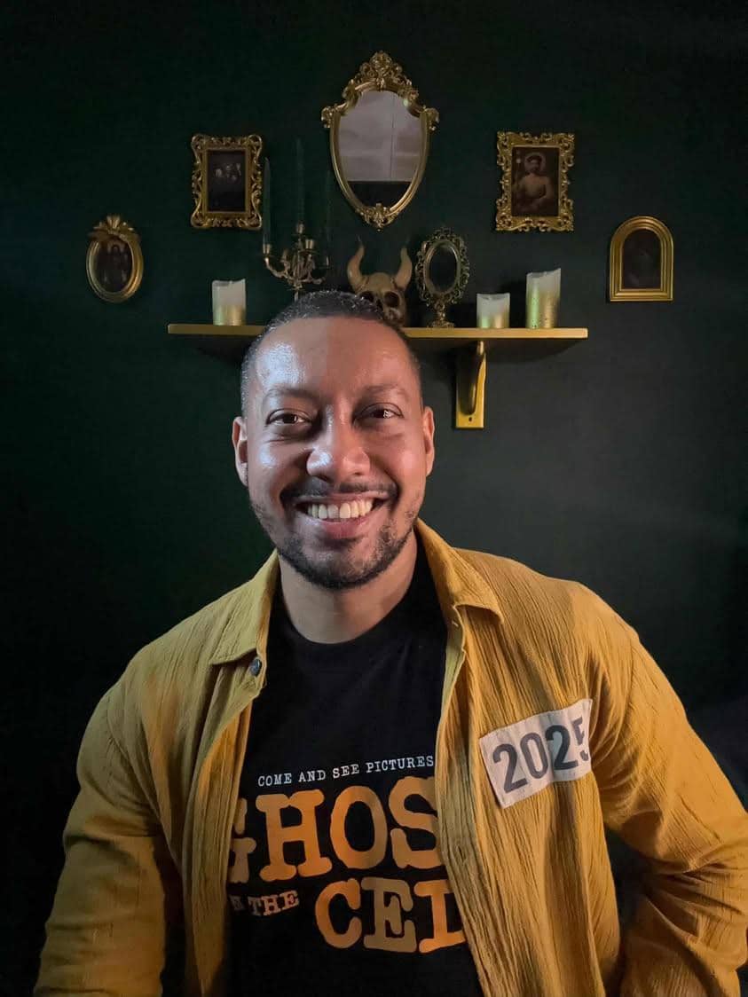 Seorang pria tersenyum mengenakan kemeja kuning dan kaus hitam bertuliskan Ghost in the Cell, berpose di depan dinding hijau dengan bingkai emas.