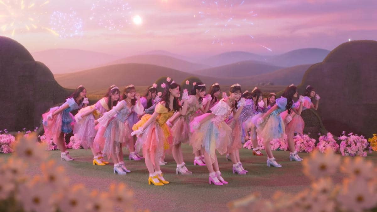 Anggota grup idola perempuan menari dengan kostum warna-warni di taman bunga dengan latar langit senja dan kembang api.