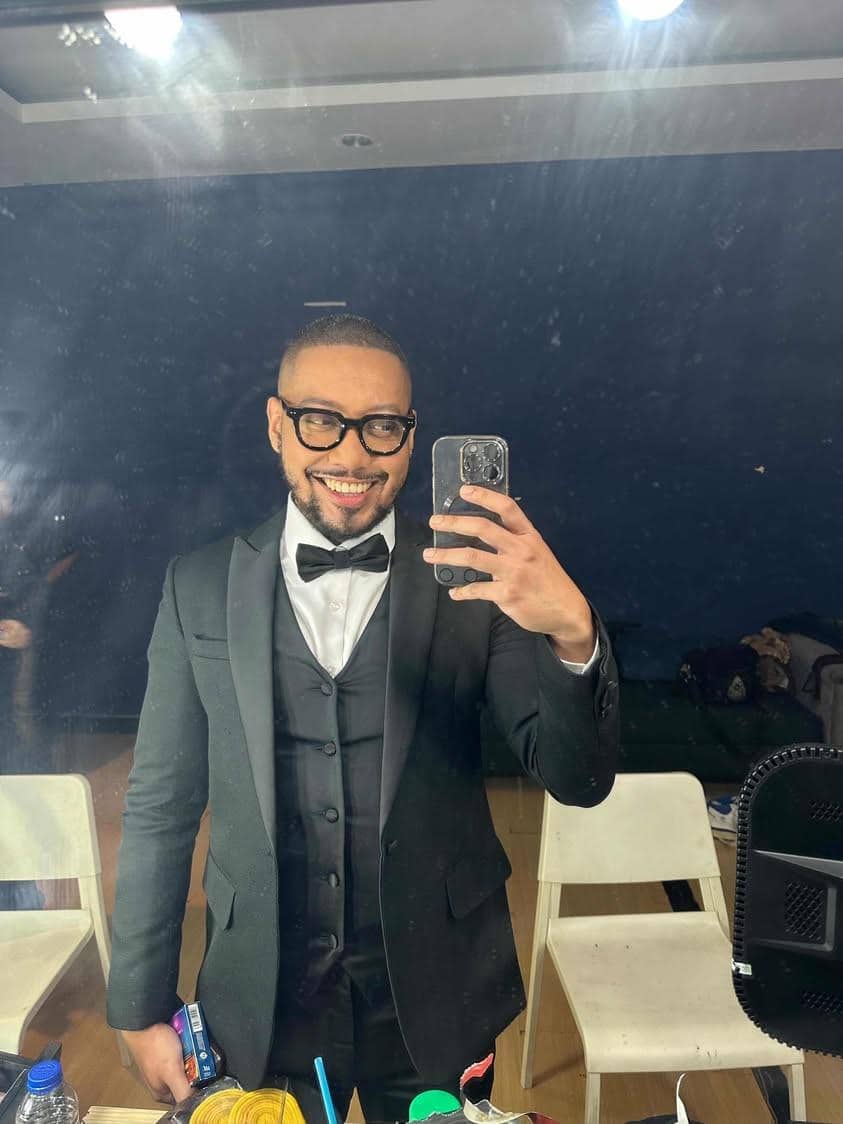 Seorang pria mengenakan setelan jas hitam dan dasi kupu-kupu tersenyum sambil berfoto selfie di depan cermin ruang rias.