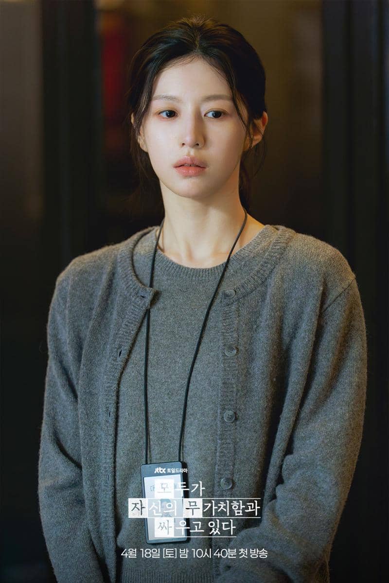 Seorang wanita berambut panjang mengenakan sweater abu-abu dan kartu identitas di lehernya dalam adegan drama We Are All Trying Here.