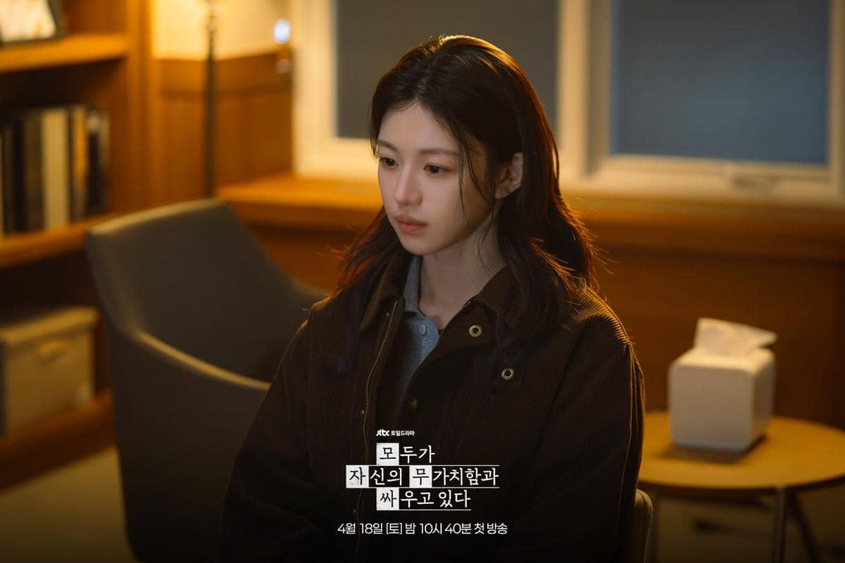 Seorang perempuan duduk di ruangan dengan pencahayaan hangat, mengenakan jaket cokelat dan tampak termenung dalam adegan drama We Are All Trying Here.