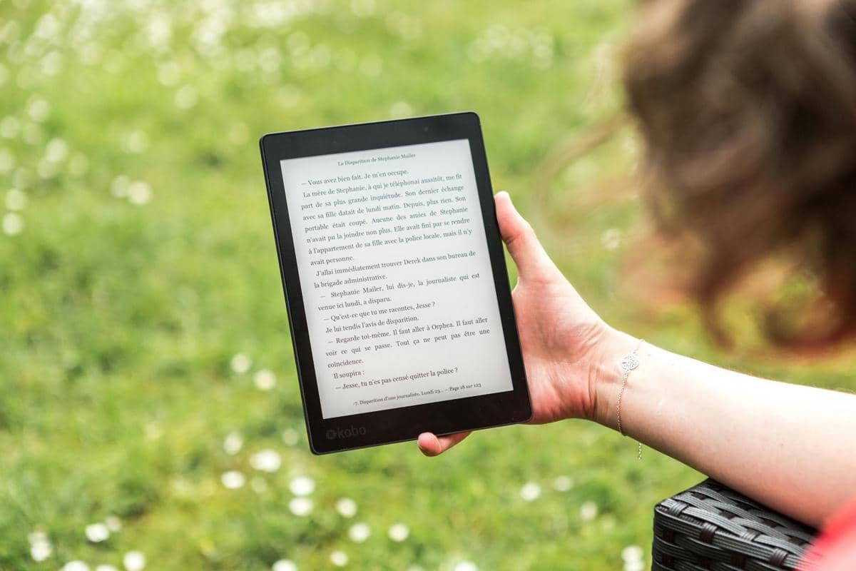 Seseorang memegang perangkat e-book hitam di luar ruangan dengan latar belakang rumput hijau dan bunga kecil yang tampak samar.