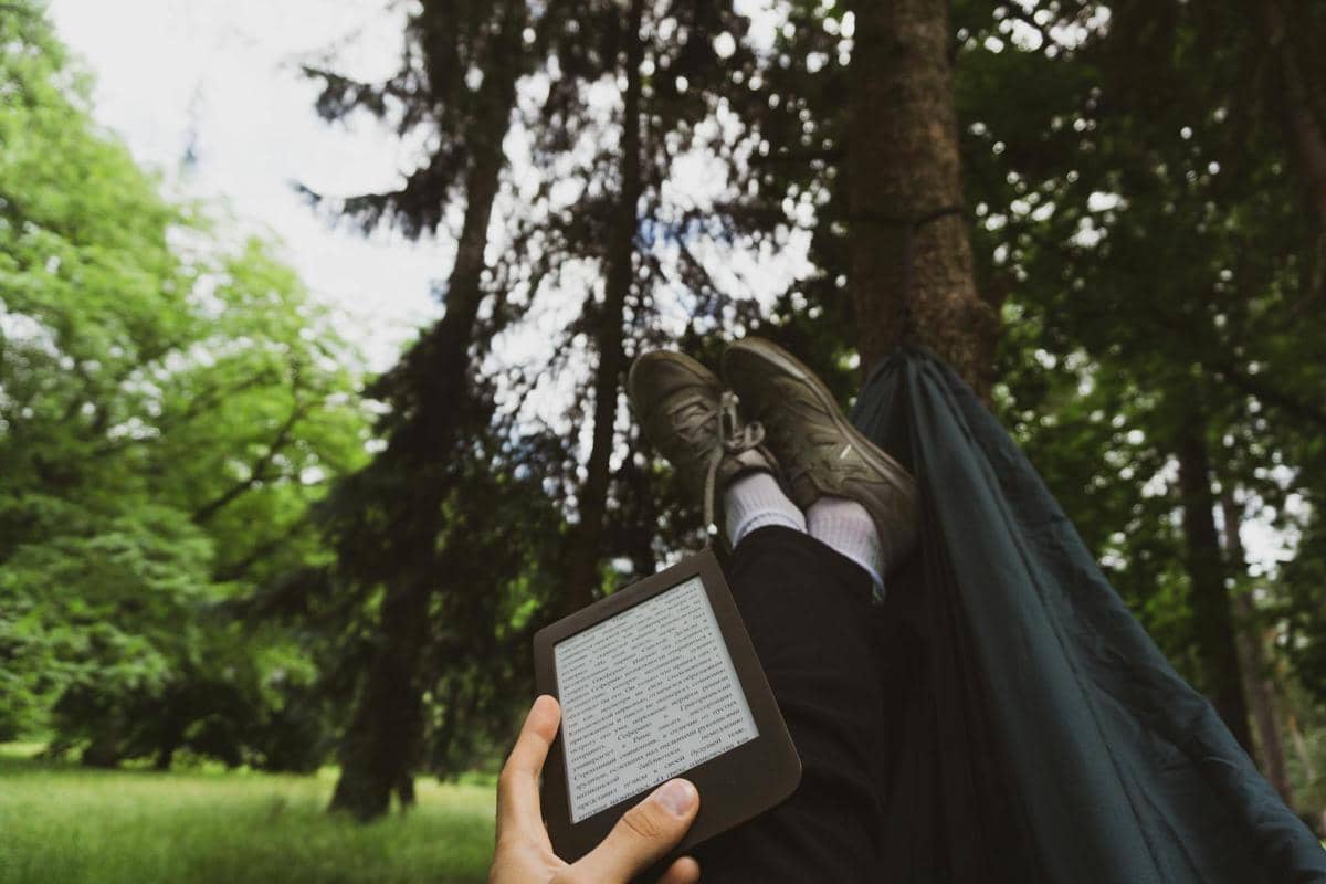 Seseorang bersantai di hammock di tengah hutan sambil membaca e-book di perangkat digital genggam dengan latar pepohonan hijau.