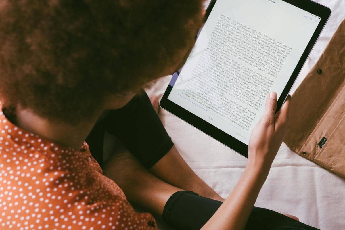 Seseorang membaca e-book di tablet sambil duduk santai di atas tempat tidur dengan pakaian kasual berwarna oranye bermotif.