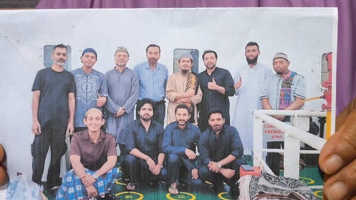 Sekelompok pria berdiri dan duduk bersama di atas kapal, mengenakan pakaian kasual dan tradisional dengan latar dek kapal berwarna putih.