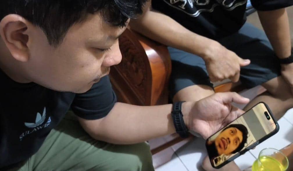 Seorang pria melakukan video call dengan seseorang di layar ponsel sambil duduk bersama rekannya di ruangan dengan pencahayaan hangat.