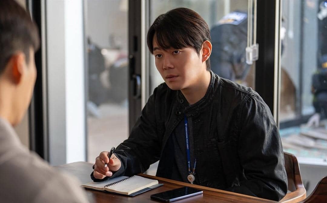 Seorang pria mengenakan jaket hitam duduk di meja dengan buku catatan dan ponsel di depannya, berbicara dengan seseorang di ruangan bercahaya alami.