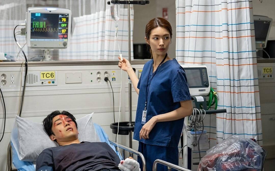 Seorang perawat mengenakan seragam biru berdiri di samping pasien pria yang terluka di ranjang rumah sakit dengan alat medis di sekitarnya.