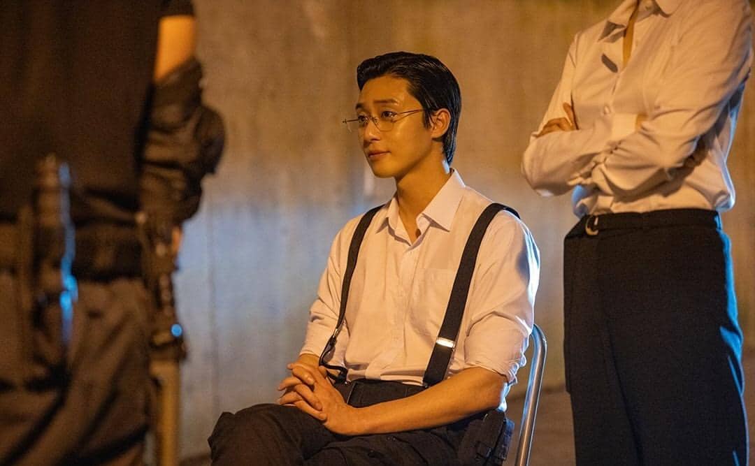 Seorang pria mengenakan kemeja putih dan suspender hitam duduk di kursi dengan ekspresi tenang di ruangan bercahaya hangat.