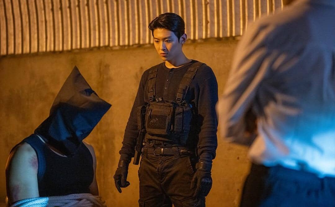 Seorang pria berpakaian hitam dengan rompi taktis berdiri di depan seseorang yang diikat dan ditutup kepalanya di tempat gelap.