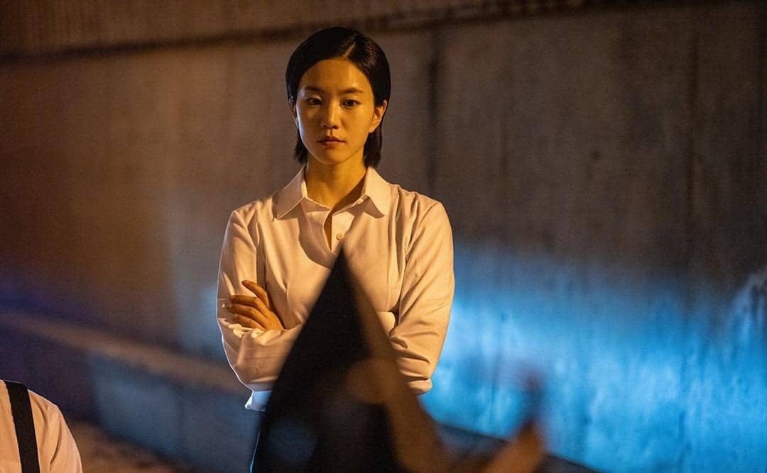 Seorang wanita berambut pendek mengenakan kemeja putih berdiri dengan tangan terlipat di depan dinding beton dengan pencahayaan malam.