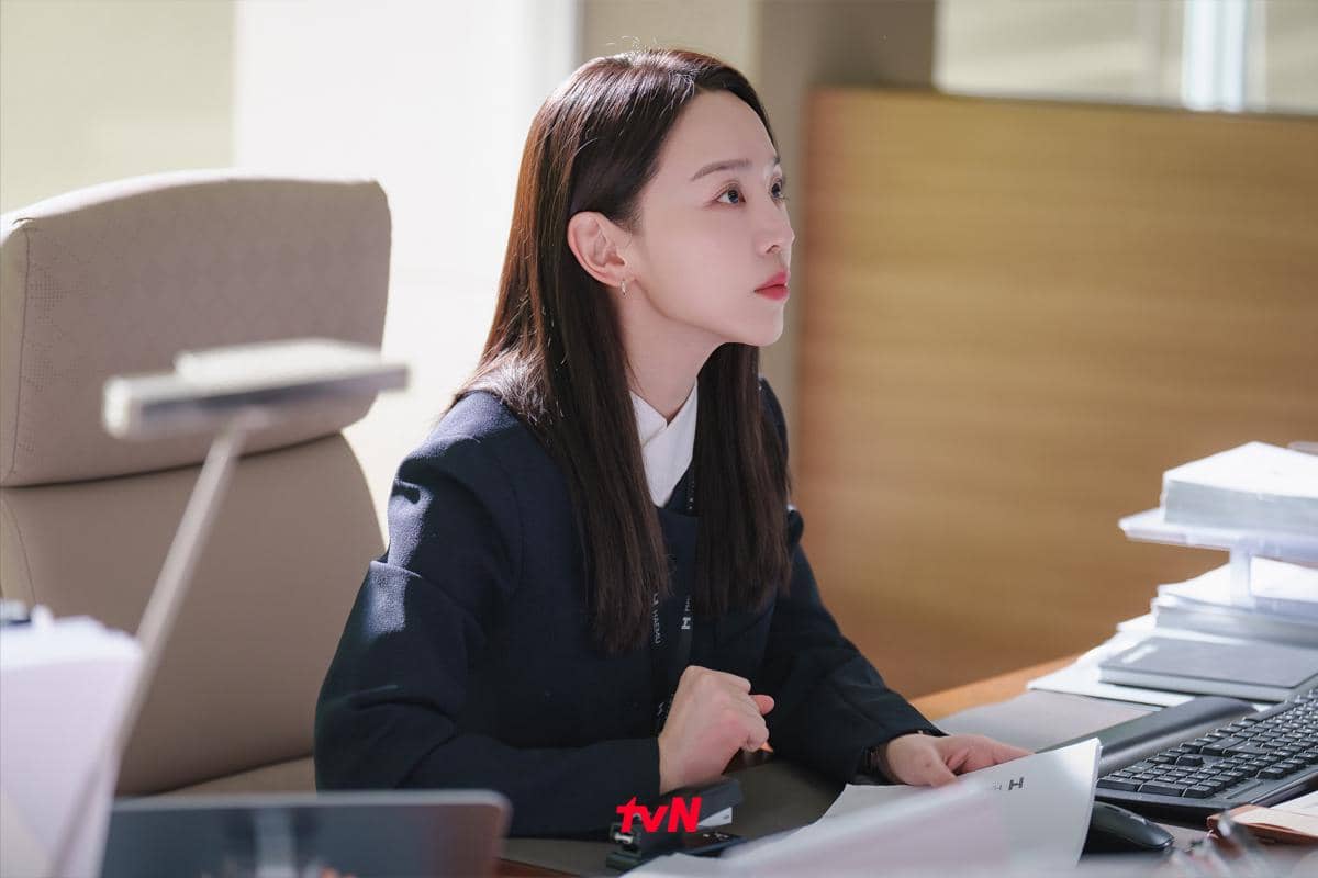 Seorang wanita mengenakan setelan kerja duduk di meja kantor dengan tumpukan dokumen dan komputer di depannya dalam adegan drama Korea Filing For Love.