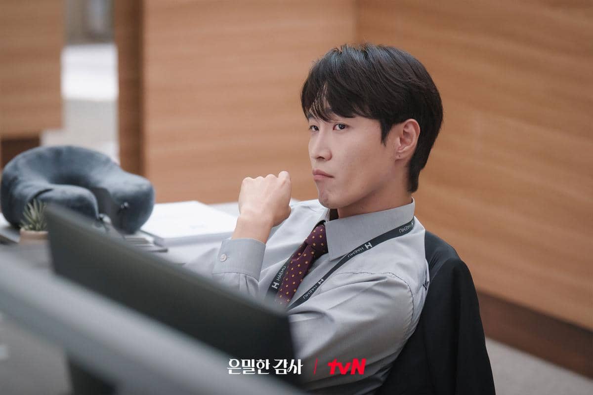 Seorang pria mengenakan kemeja abu-abu dan dasi bermotif duduk di meja kantor dengan ekspresi serius dalam cuplikan drama Korea Filing For Love.