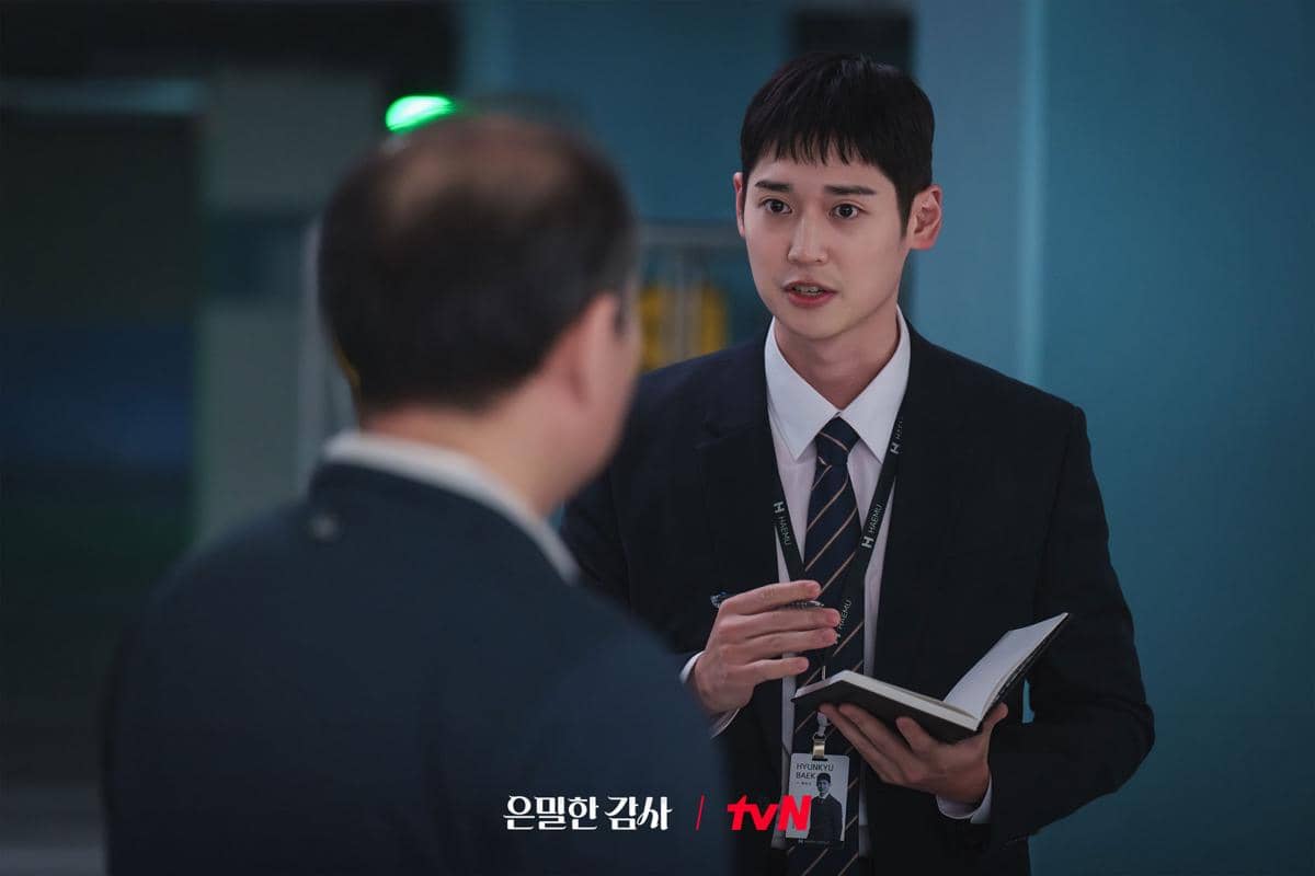 Seorang pria mengenakan setelan jas dan dasi sedang berbicara sambil memegang buku catatan di hadapan pria lain di ruangan kantor.