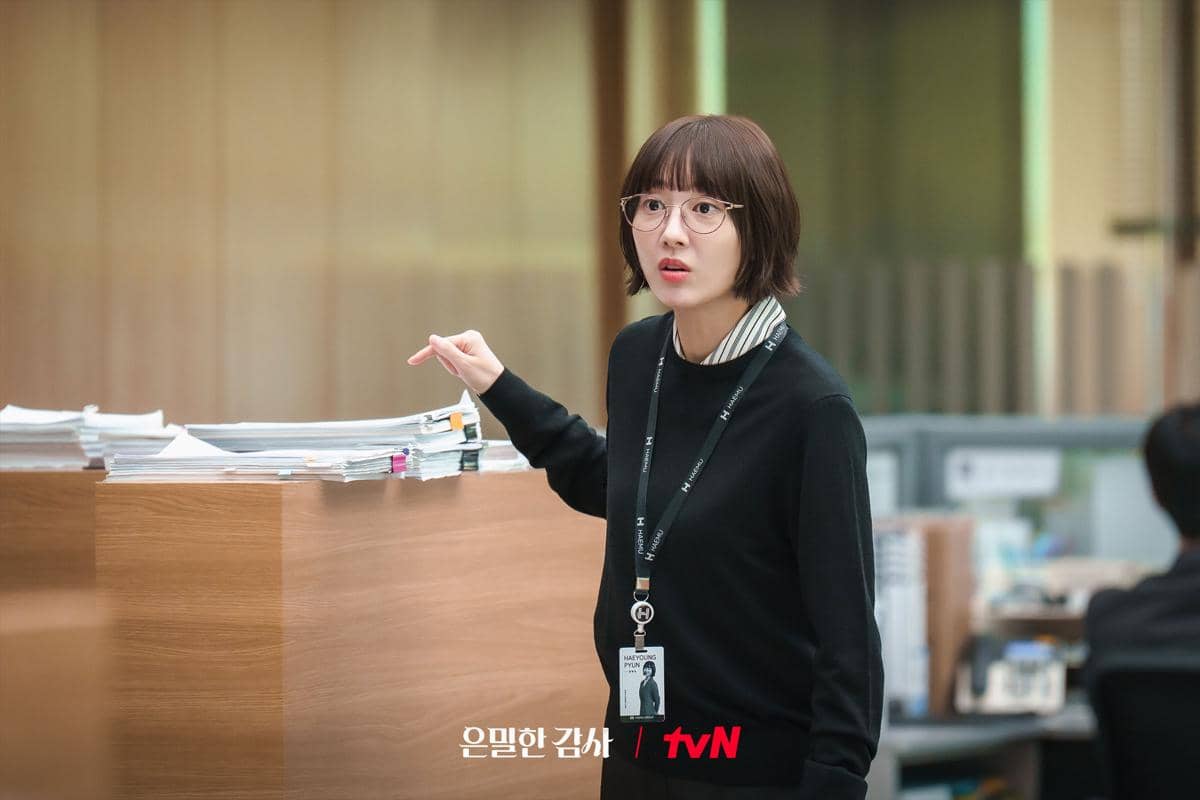 Seorang wanita berambut pendek mengenakan kacamata dan pakaian kantor hitam berdiri di dekat tumpukan berkas di meja kerja.