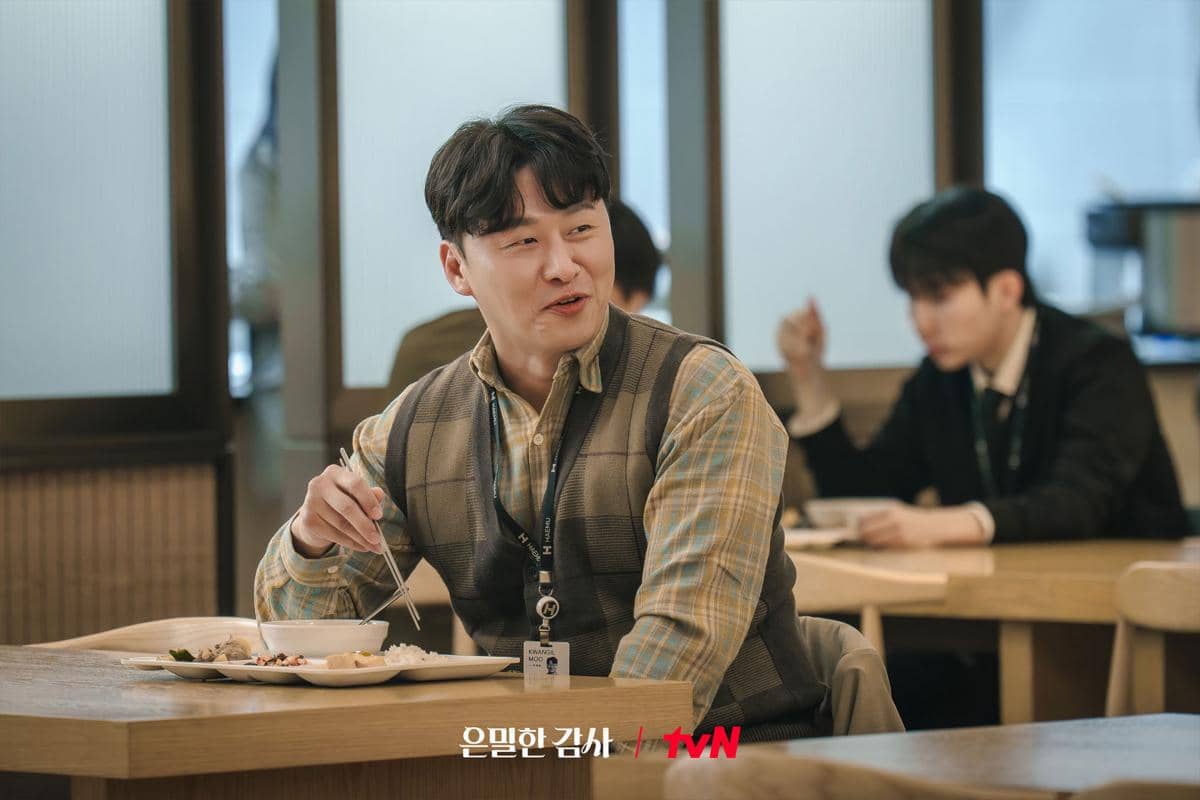 Seorang pria mengenakan kemeja kotak dan rompi duduk di kantin sambil makan dan berbicara, dengan suasana hangat di sekitarnya.