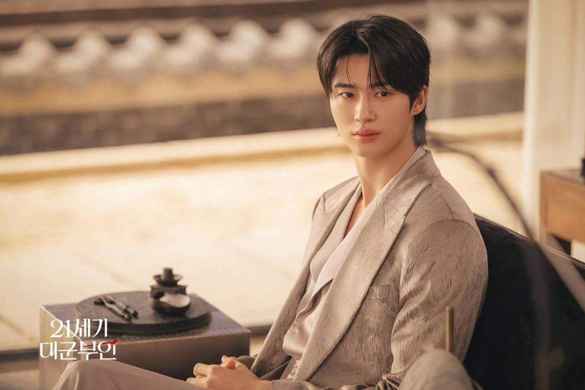 Seorang pria mengenakan setelan jas berwarna krem duduk di dalam ruangan bergaya tradisional dalam cuplikan drama Korea Perfect Crown.