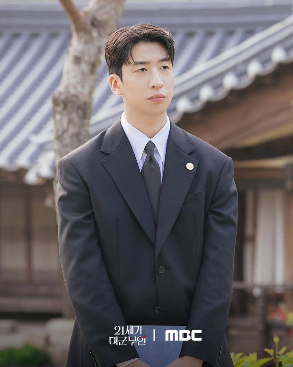 Seorang pria mengenakan setelan jas hitam berdasi berdiri di luar bangunan tradisional Korea dalam cuplikan drama Perfect Crown.