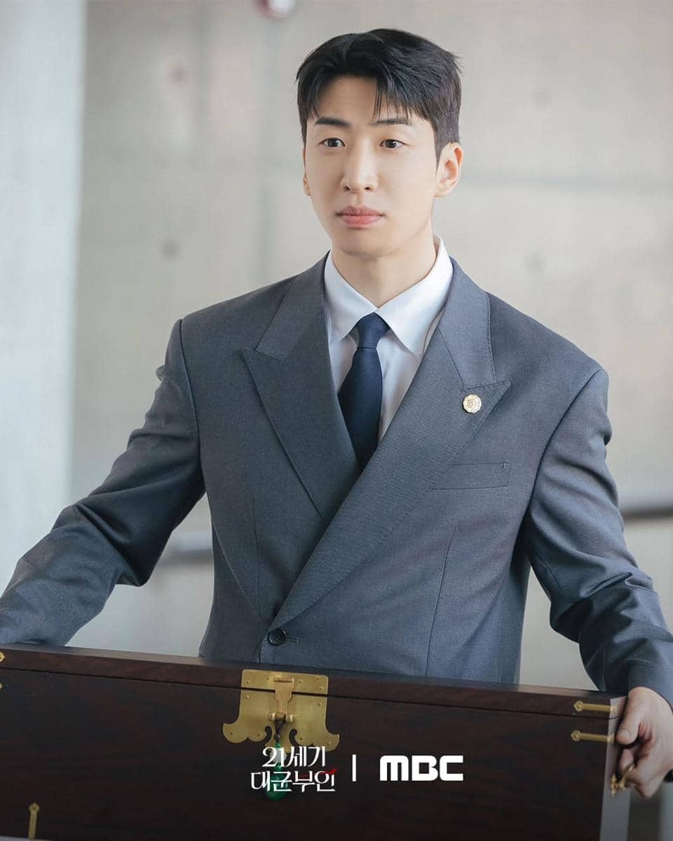 Seorang pria mengenakan setelan jas abu-abu dan dasi biru tua berdiri sambil memegang kotak kayu besar dengan latar ruangan modern.