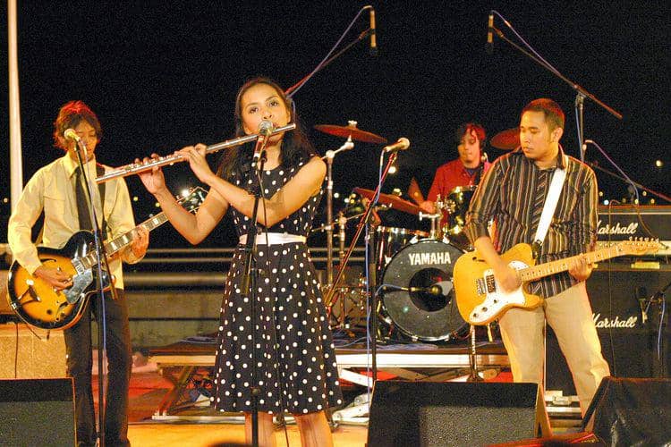 Grup musik Mocca sedang tampil di panggung pada malam hari; Arina Ephipania sambil meniup flute, diiringi oleh pemain gitar, bass, dan drum di latar belakang.