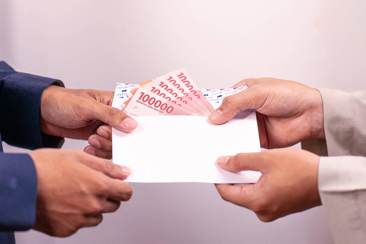 Dua orang saling menyerahkan amplop berisi uang pecahan seratus ribu rupiah sebagai ilustrasi pembayaran upah pekerja rumah tangga.