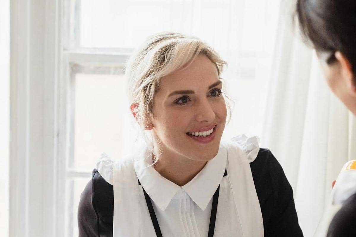 Seorang perempuan mengenakan seragam hitam putih tersenyum ramah saat berbicara dengan seseorang di ruangan bercahaya alami.