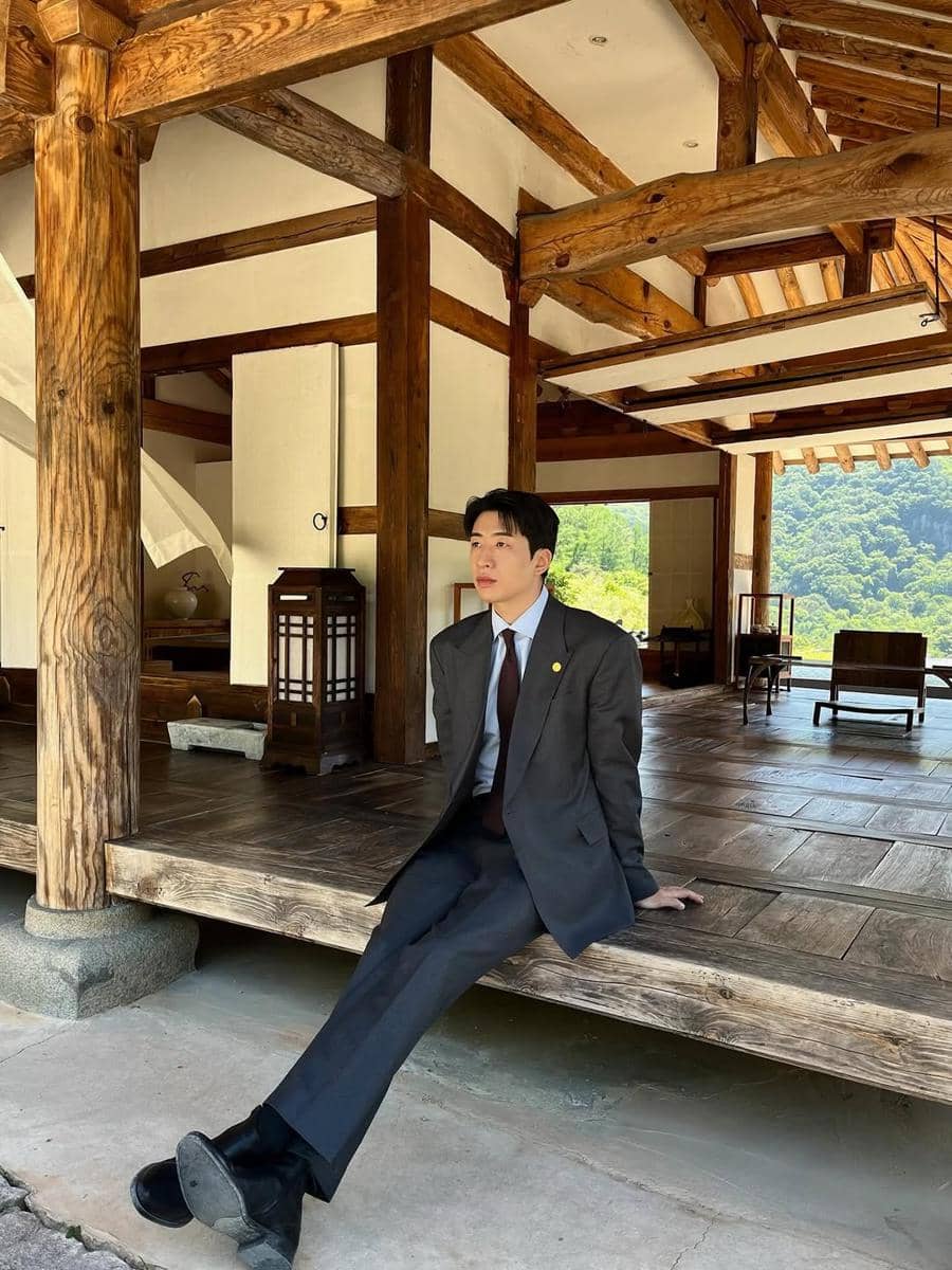 Seorang pria mengenakan setelan jas duduk di teras bangunan kayu tradisional dengan latar pemandangan hijau pegunungan.