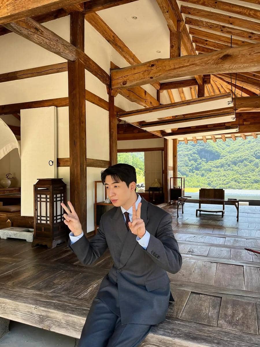 Seorang pria mengenakan setelan jas abu-abu duduk di teras bangunan kayu tradisional sambil berpose dengan dua tanda damai di tangannya.