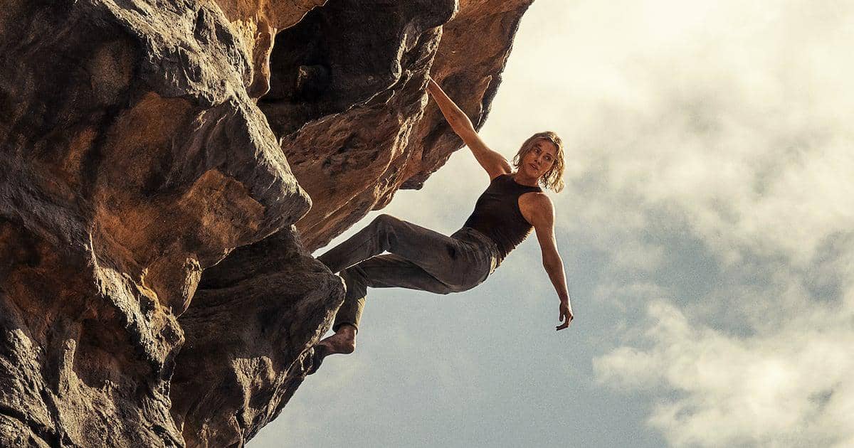Seorang perempuan memanjat tebing batu besar dengan tangan menggantung di ketinggian di bawah langit cerah dalam adegan film Apex.