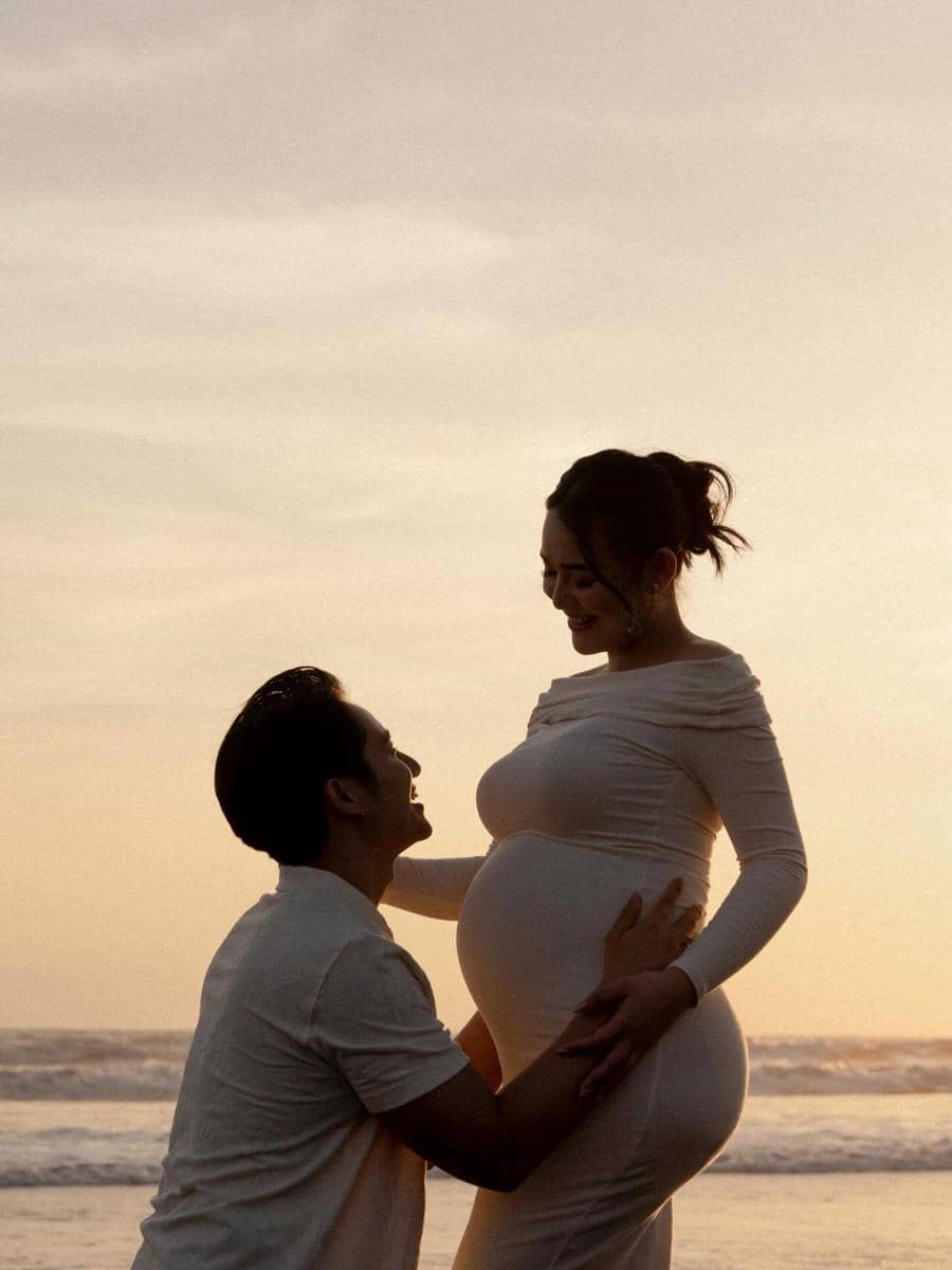 Seorang wanita hamil mengenakan gaun putih berdiri di tepi pantai saat matahari terbenam, sementara seorang pria berlutut di depannya dengan ekspresi bahagia.