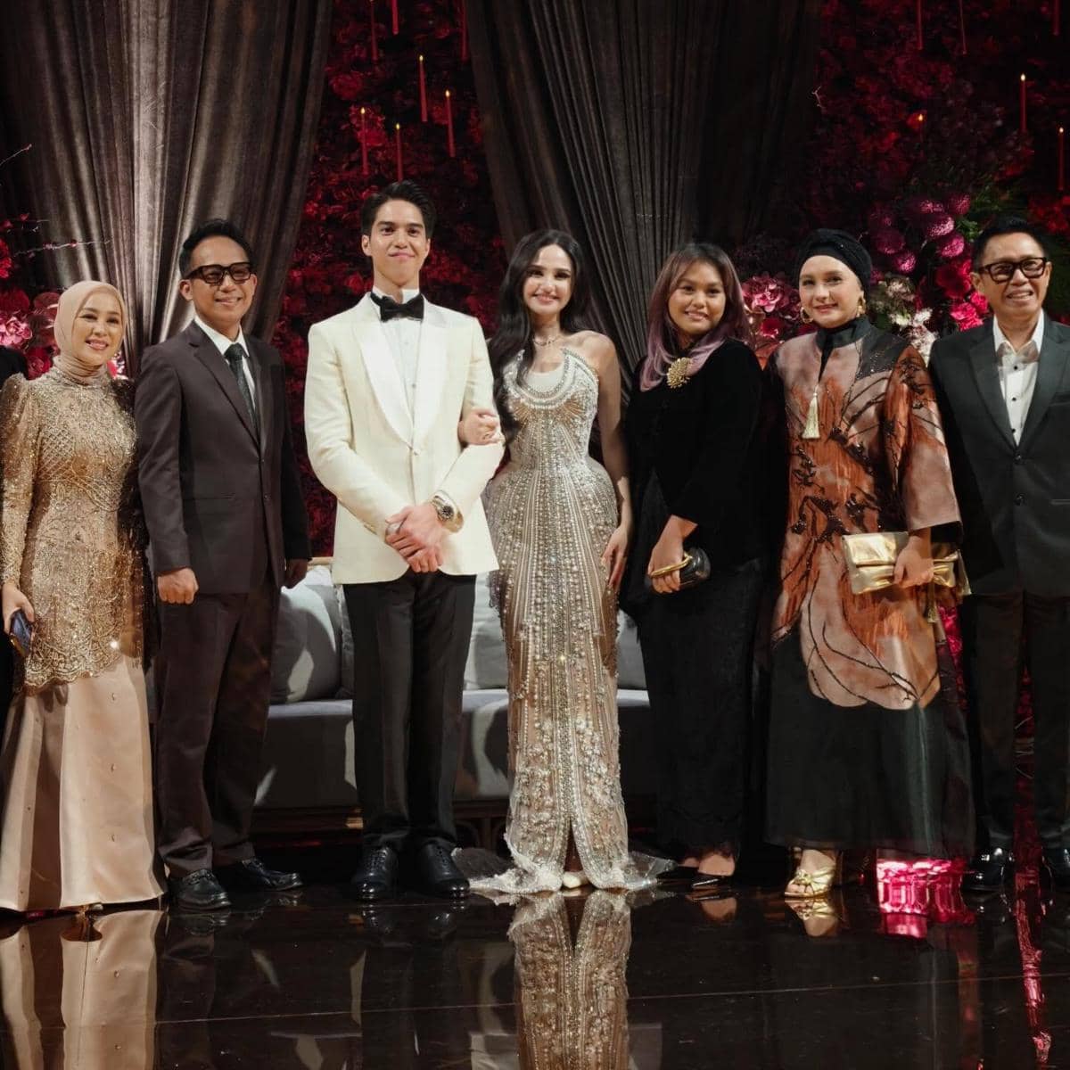 Beberapa tamu mengenakan busana formal berpose bersama pasangan pengantin di resepsi pernikahan dengan latar dekorasi bunga merah.