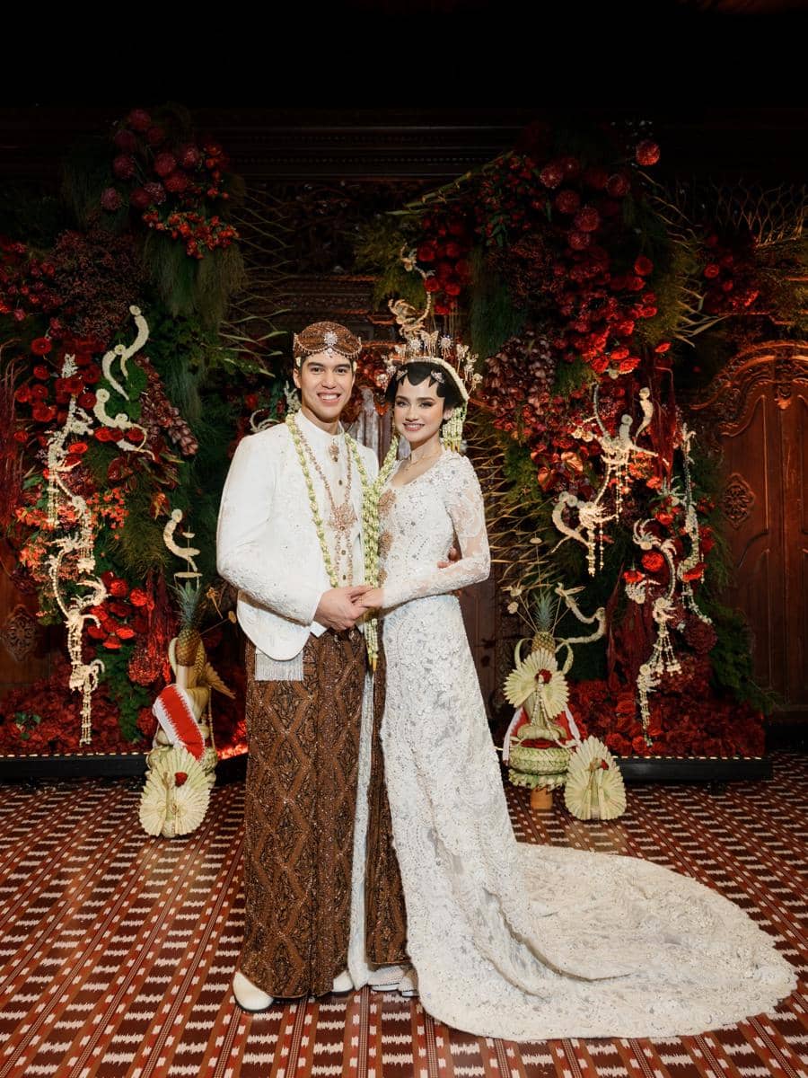 Sepasang pengantin mengenakan busana adat Jawa berwarna putih dan cokelat berpose di depan dekorasi bunga merah dan hijau yang megah.