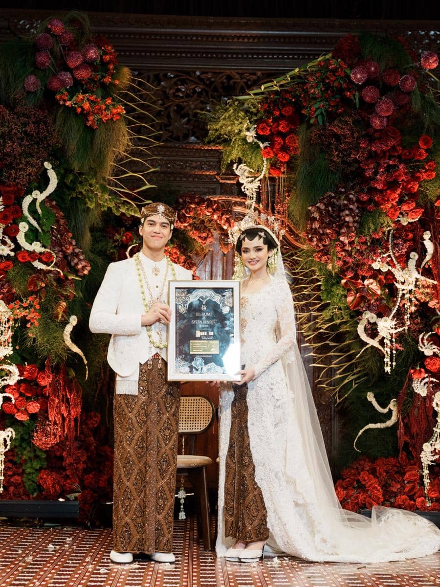 Sepasang pengantin mengenakan busana adat Jawa berwarna putih dan batik cokelat berpose sambil memegang bingkai sertifikat di pelaminan berhias bunga merah.