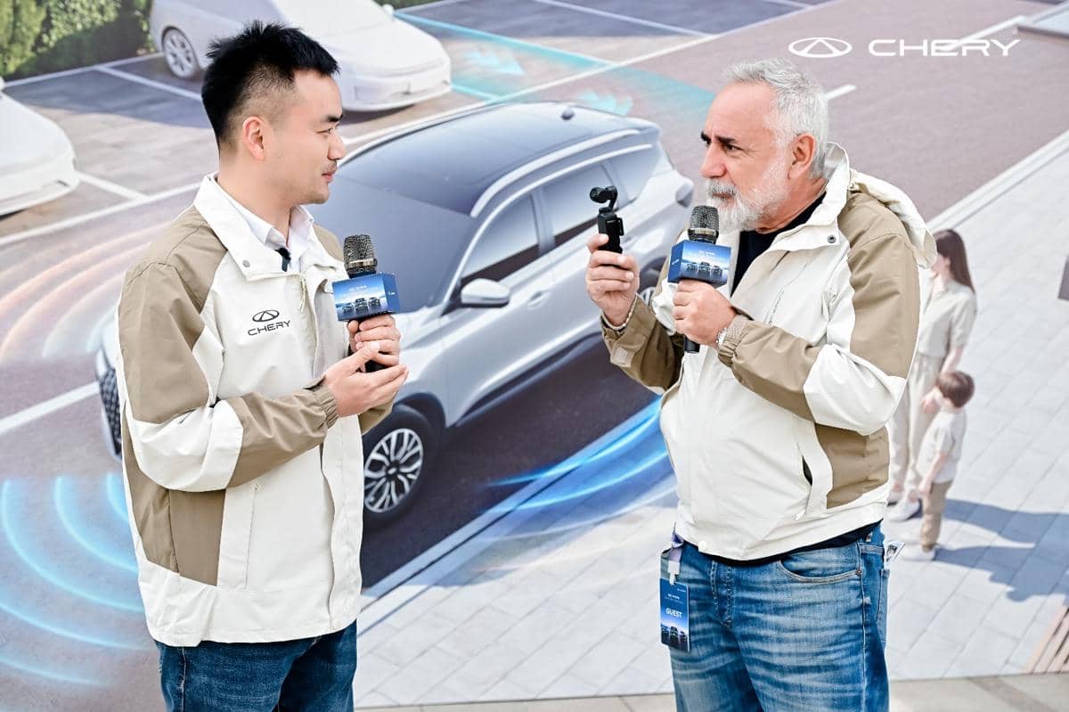 Dua pria mengenakan jaket Chery berbicara sambil memegang mikrofon di depan latar mobil dengan logo Chery dalam acara demonstrasi global.