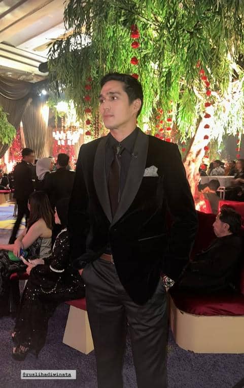 Seorang pria mengenakan setelan jas hitam berdasi berdiri di area resepsi pernikahan dengan dekorasi pohon dan lampu mewah di sekitarnya.